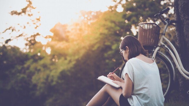 A person sitting on grass, writing in a notebook with a pen on a creative writing retreat. They're facing away, wearing glasses and a white shirt. A bicycle with a wicker basket is nearby. Sunlight filters through trees in the background.
