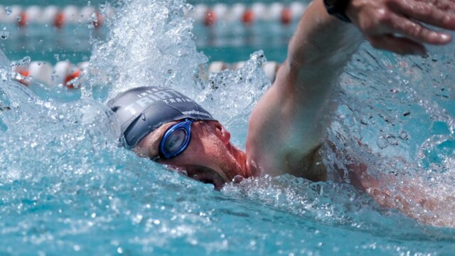 A swimmer wearing goggles and a swim cap performs a freestyle stroke in a pool on the Swim Well camp. Splashes of water surround their face and arm, which is extended forward. The pool lanes are visible in the background.
