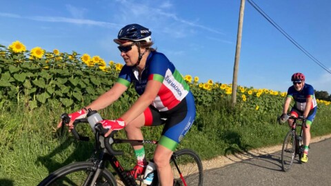 Two cyclists ride on a country road bordered by sunflowers under a clear blue sky. They wear matching blue, green, red, and white cycling jerseys. The lead cyclist smiles, while the second cyclist follows closely behind.