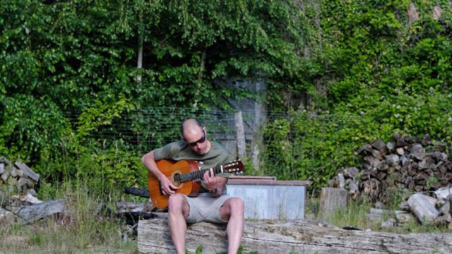 A person in sunglasses, wearing a green shirt and shorts, plays an acoustic guitar while sitting on a log in a grassy area. Behind them is lush greenery and stacked logs, creating a serene outdoor vibe for the Make Music Together experience.