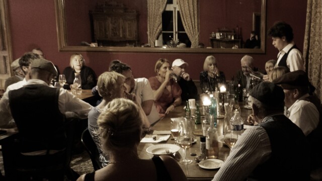 People gather around warmly lit dining tables, engaged in conversation, as the Death at the Chateau, immersive murder mystery unfolds. The rooms red walls and curtained windows add to the ambiance. Candles and drinks on the table create a warm, convivial atmosphere.