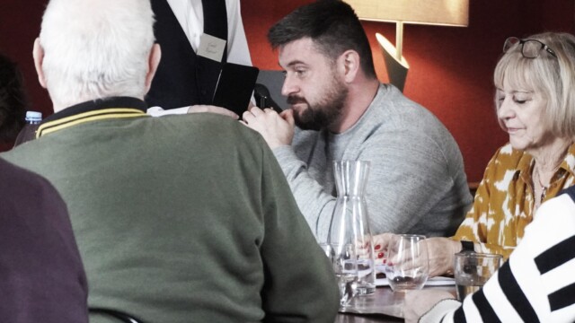 A group of people sit around a dining table, engaging in our Murder Mystery. The young detective stands by as a man in a grey sweater speaks with him. Glasses and a water carafe adorn the table, while a warm lamp glows in the background.