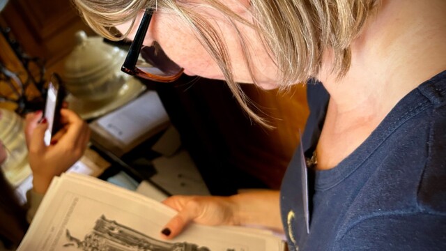A short blond-haired woman with glasses examines an open book, showcasing ornate vases and furniture illustrations from the Death at the Chateau, an immersive Murder Mystery weekend.