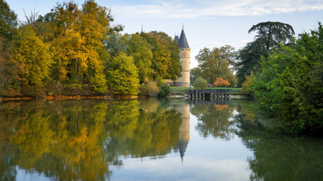 A serene landscape with a tower surrounded by autumn trees, reflected in a tranquil lake. The sky is partly cloudy, adding depth to the peaceful scene. A wooden pier extends into the water, enhancing the rustic charm.