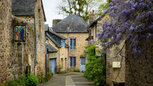 Narrow stone-paved alley in a quaint village with rustic stone houses. Purple wisteria cascades from the right. A colorful, medieval-style sign hangs on the left. Lush greenery lines the pathway under a cloudy sky.