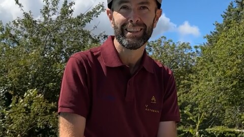 Joel Filliol, wearing a maroon polo shirt and a black cap, sits on a low wall by the pool at the Joel Filliol Retreat, smiling at the camera against a backdrop of trees and a blue sky with clouds.