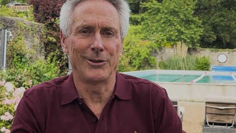 Mark Allen, wearing a maroon polo from the Mark Allen Experience training camp sits by the pool, with green foliage and a stone wall in the background.