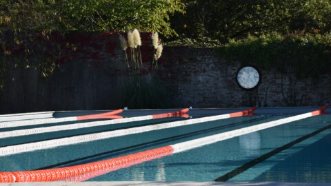 Outdoor swimming pool with clear water, red and white lane dividers, a wall covered in green and red plants, and a large round clock on the wall. Trees and bushes surround the area—perfect for a run training retreat with Nick Bester.