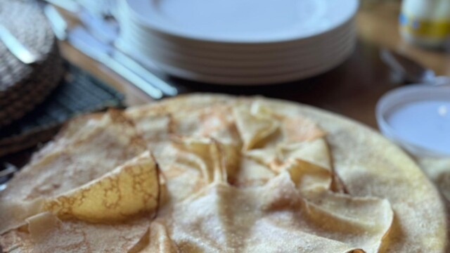 A plate of thin, golden crepes sits on a wooden table set for a meal at a glorious chateau in the Loire, with stacked white plates, cups, cutlery, and a fruit basket visible in the background.