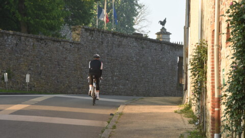 A cyclist wearing a helmet and cycling gear rides on a quiet road next to a stone wall with flags and a rooster statue visible in the background. Sunlight casts long shadows along the path.