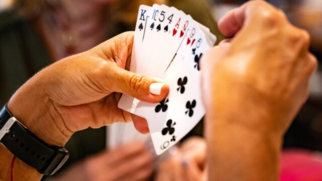 A person holds a fan of playing cards, mostly clubs and one heart, while another person sits across the table with blurred hands and face in the background. The focus is on the cards and hands.