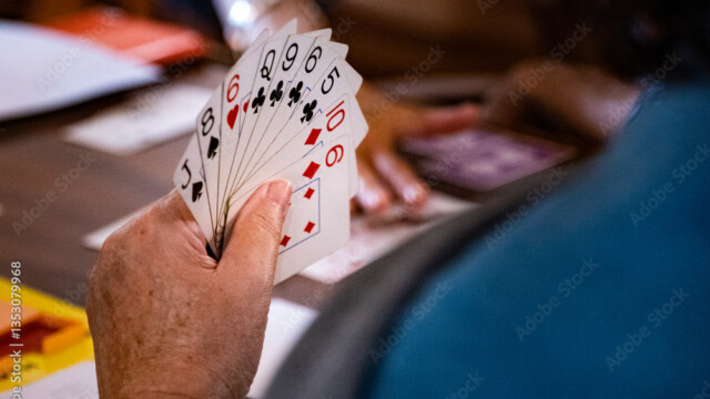 A person holds a fan of playing cards, including the 2, 5, 6, 9, and 10 of clubs, and the 10 of hearts, with another person and cards visible in the blurred background.
