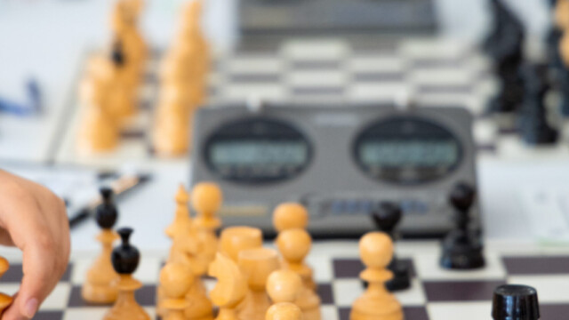 Close-up of a chess game in progress, with a childs hand moving a piece. Multiple chessboards and digital timers are visible in the background, and a black chess piece with a red sticker is tipped over on the board.