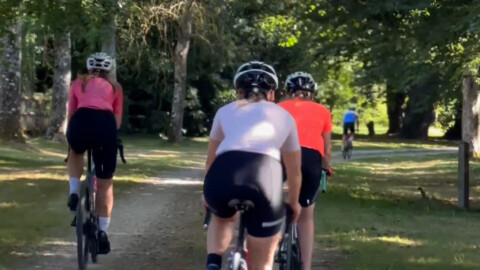 A group of cyclists wearing helmets and colorful jerseys ride along a tree-lined gravel path in a park on a sunny day.