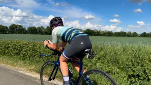 A cyclist wearing a helmet and cycling gear rides a road bike alongside a green field under a blue sky with scattered clouds. The rider is leaning forward, pedaling on a sunny day.