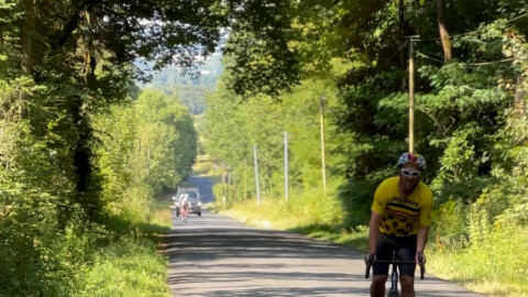 A cyclist in a yellow jersey rides uphill on a tree-lined road in sunlight, with several more cyclists visible in the distance behind him.