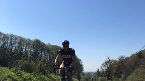 A cyclist wearing a helmet and cycling gear rides on a sunny rural road, with trees lining the roadside and another cyclist visible in the distance under a clear blue sky.