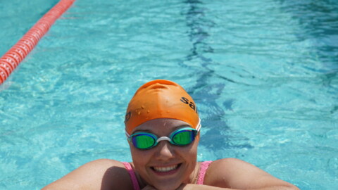 A smiling swimmer wearing an orange swim cap and green goggles rests their arms on the edge of an outdoor pool, with clear blue water and lane dividers visible in the background.