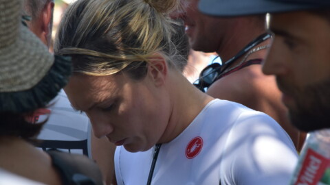A woman in a white athletic outfit with a red logo looks down, surrounded by people outdoors near a glorious chateau in the Loire. The image is candid and focused on her, with blurred individuals in the foreground and background.