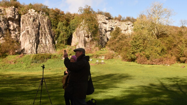 A man in a flat cap takes a photo with his phone in a grassy field, beside a tripod. Behind him are rocky cliffs, trees, and a picnic table—reminiscent of the picturesque grounds near a glorious chateau in the Loire.