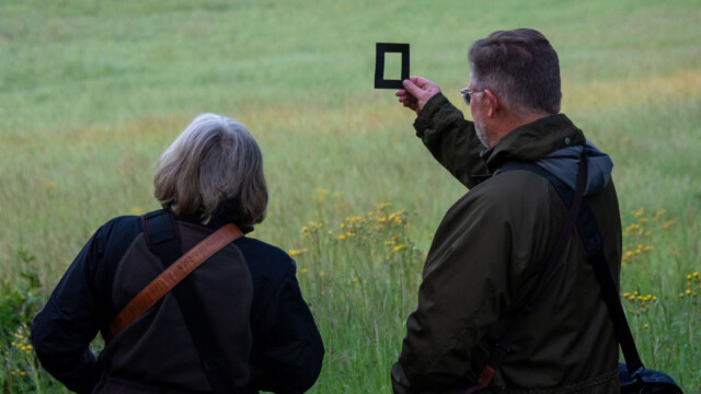 Two people with shoulder bags stand in a grassy field near a glorious chateau in the Loire. One person holds up a small rectangular black frame, looking through it towards the landscape. Both are facing away from the camera.