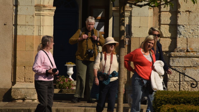 Five women with cameras stand and walk outside a glorious chateau in the Loire on a sunny day. Some wear hats and sunglasses, while one woman in the foreground has a coral sweater and white jacket. Greenery and a tree are nearby.
