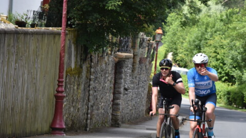Two cyclists ride on a narrow road next to a stone wall and greenery. Both are wearing helmets and sunglasses; one in black, one in blue. They are giving thumbs up and appear to be enjoying their ride.