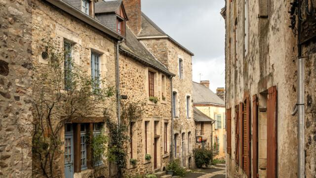A narrow cobblestone street lined with old stone buildings and shuttered windows evokes the charm of a glorious chateau in the Loire. A wrought-iron sign reading Aubabite Medievale hangs above, beneath an overcast sky in this quaint village.