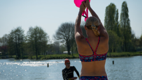 A woman in a colorful swimsuit stands in shallow lake water, holding a bright pink inflatable above her head, while another person in a black wetsuit approaches from behind. Trees line the background under a clear sky.