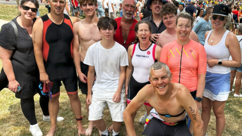 A group of smiling people, some in triathlon gear and some in casual clothes, pose together outdoors on grass at a busy event with a crowd and finish line in the background.