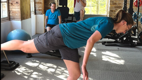 A woman in athletic clothes balances on one leg with her arms extended forward at a gym. Two other people are in the background, one sitting and one standing, near exercise equipment and large windows.