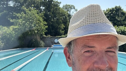 A man wearing a light-colored hat and navy shirt smiles at the camera in front of an outdoor swimming pool with lane dividers, surrounded by trees and greenery under a clear blue sky.