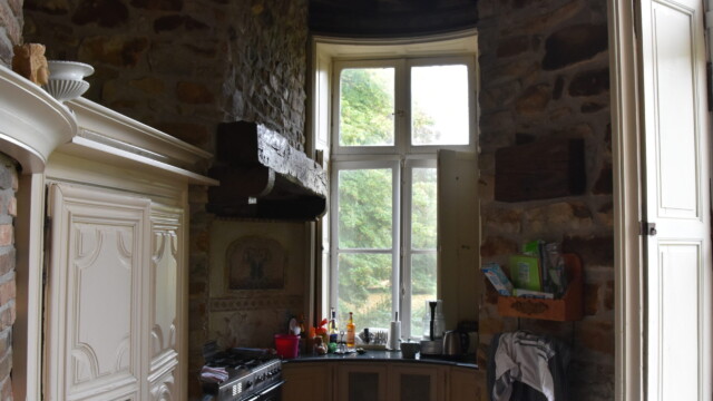 A rustic kitchen with stone walls, a large window letting in daylight, wooden shelves, a black oven, various bottles and kitchen items on the counter, and cream-colored cabinets.