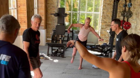 Four adults exercise together in a gym with stone walls and large windows. Two people are balancing on one leg, while others stand nearby, engaged in the activity. Fitness equipment is visible in the background.