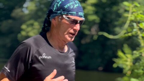 A man wearing sunglasses, a headscarf, and a black parkrun shirt runs outdoors on a sunny day, with trees and greenery blurred in the background.