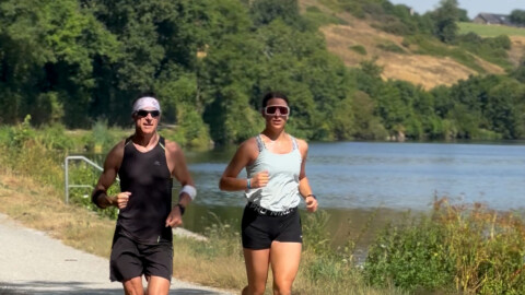 Two people wearing athletic clothes and sunglasses are jogging on a riverside path surrounded by greenery and hills on a sunny day. A calm river is visible to their right.
