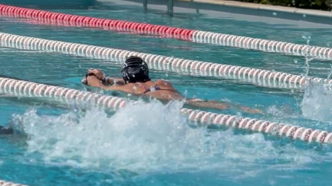A swimmer in a pool wearing a swim cap and goggles performs the breaststroke in a lane, while water splashes nearby, possibly from another swimmer. The pool is divided by red and white lane dividers.