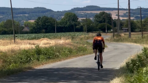 A cyclist wearing an orange and black outfit rides alone on a narrow rural road, surrounded by fields, greenery, and distant hills under a partly cloudy sky.