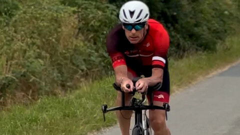 A cyclist in a red and black outfit, wearing a white helmet and sunglasses, rides a road bike on a rural lane bordered by green bushes and grass.