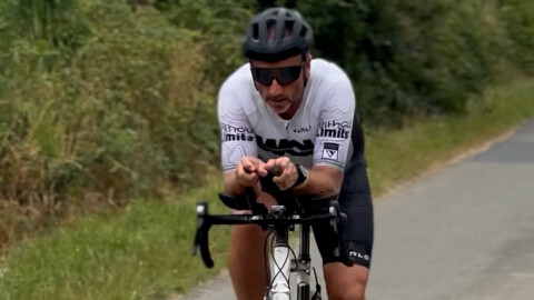 A cyclist wearing a helmet, sunglasses, and a white jersey rides on a road bike in an aerodynamic position, with trees and greenery in the background.