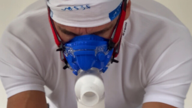A person wearing a white shirt, headband, and a blue respiratory face mask is focused downward, possibly engaged in a physical or medical activity.