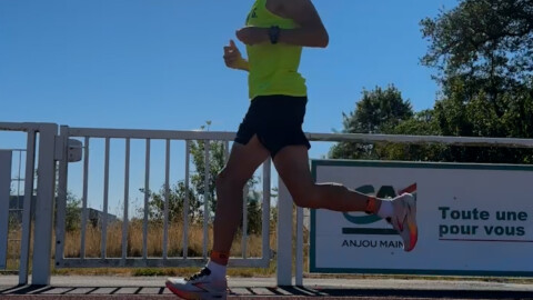 A person in a bright yellow tank top and black shorts is running outdoors near a white fence, with trees and a clear blue sky in the background.