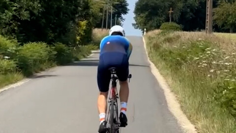 A cyclist in a blue outfit and white helmet rides a road bike uphill on a rural, tree-lined road under a clear sky.