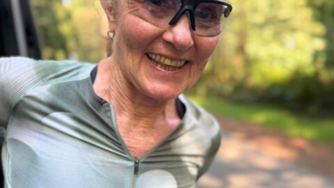 An older woman with light hair tied back smiles outdoors. She wears sporty sunglasses and a zipped-up athletic top. The background is a sunny, tree-lined path in a park or forest.
