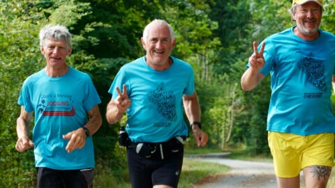 Three older men jog on a tree-lined gravel path. All wear blue shirts, and one wears yellow shorts. They smile and make peace signs, appearing cheerful and energetic during their run outdoors.