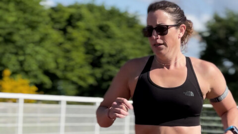 A woman wearing sunglasses, a black sports bra, and an armband runs outdoors on a sunny day, with green trees and a fence in the blurred background.