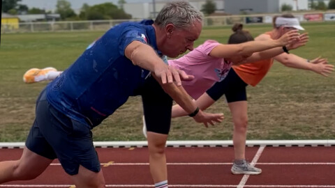 A group of people stretch their arms and legs forward in a balancing pose on a running track, with one man in front wearing a blue shirt and others behind him in pink and orange shirts.