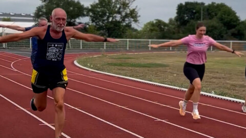 An older man and a woman run on an outdoor track with their arms outstretched like airplane wings, both smiling and having fun. Trees and fencing are visible in the background.