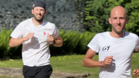 Two men in white t-shirts are jogging outdoors on a sunny day. One is in the foreground smiling, while the other is slightly behind and appears focused. Greenery and a stone wall are visible in the background.