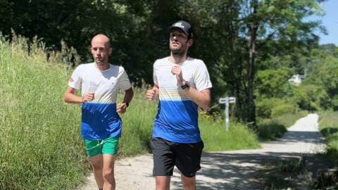 Two men are jogging on a gravel path surrounded by greenery and trees. Both wear matching white shirts with blue and green stripes; one wears green shorts, the other black shorts and a black cap. Sunlight filters through the trees.